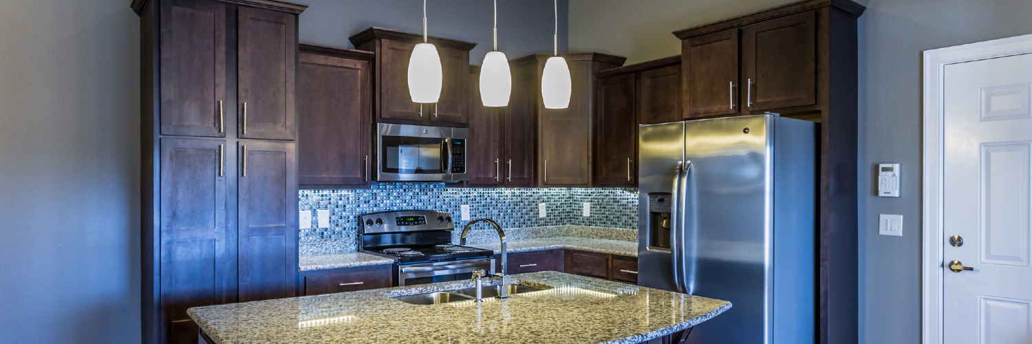 Photo of an empty kitchen with stainless-steel appliances, granite counter tops, custom dark wood cabinetry, and a blue glass backsplash. It's such a luxurious kitchen for a one-bedroom apartment in Little Rock.