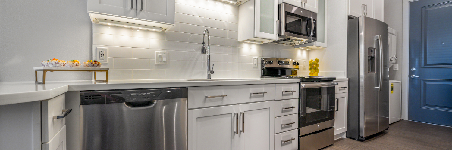 Photo of a luxury apartment kitchen in a studio at The Pointe North Hills in North Little Rock, Arkansas. A white nano stone counter top, stainless steel appliances, subway tile backsplash, gooseneck faucet, stacked washer/dryer unit, and white cabinets are showing.