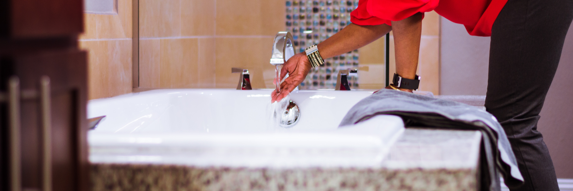 Photo of woman in red shirt starting a bath in a large soaking tub in a bathroom. She is wearing a metallic bracelet and a watch. There is a gray towel on the bath ledge.