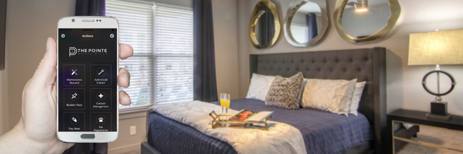 Photo of a hand holding a cell phone showing The Pointe North Hills resident app. In the background, there is a bedroom set in a luxury apartment at The Pointe North Hills in North Little Rock, Arkansas.