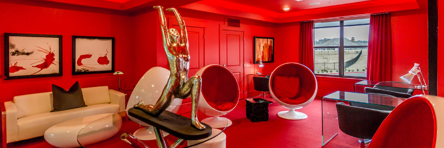Photo of the Zen Lounge inside the Bowman Pointe community clubhouse in Little Rock, Arkansas. The room has red walls, floors, doors, curtains, and reading pods. There is also a white couch and glass desks. You can see a silver abstract sculpture of a person doing yoga on a glass table.